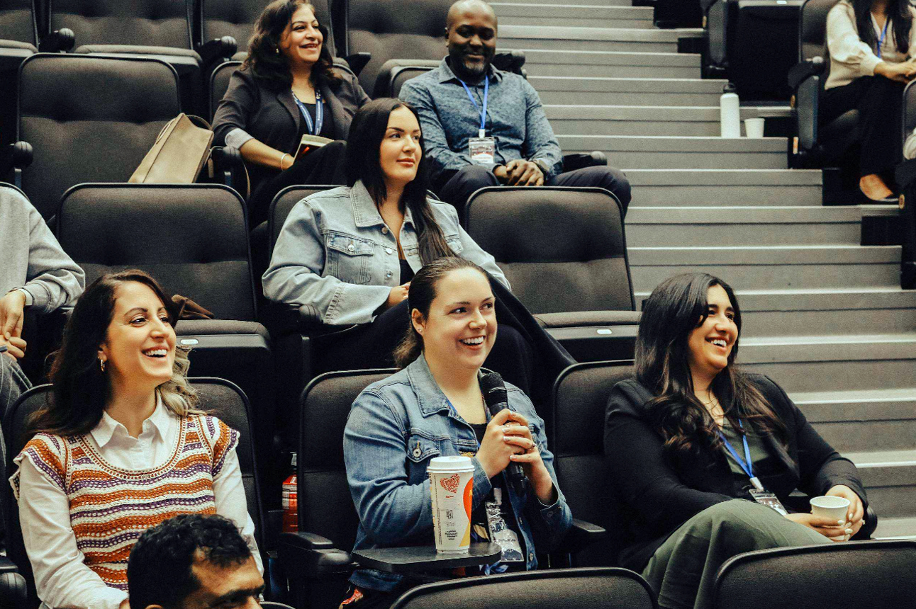 Participants sit in a tiered lecture hall, smiling and listening during an interactive presentation. One attendee in the front row holds a microphone while speaking, as others around them laugh and engage in the discussion. Coffee cups, reusable water bottles, and conference name badges are visible, reflecting a welcoming and collaborative professional development session.