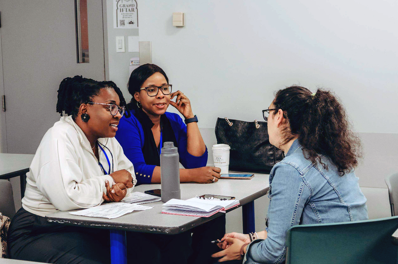 Three conference participants sit around a table engaged in a small-group discussion. Two participants face the camera while listening and speaking with a third participant seated with their back to the camera. Notebooks, papers, reusable water bottles, and coffee cups are on the table, reflecting a collaborative professional development session focused on conversation and idea sharing.