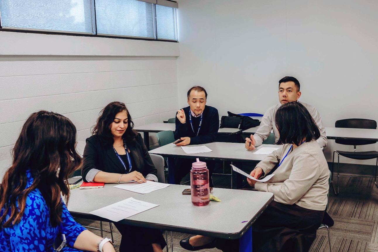 Five conference participants sit around tables in a bright classroom, engaged in a small-group discussion. Several participants hold printed handouts while one person gestures as they share an idea. Notebooks, reusable water bottles, and mobile phones are on the tables, highlighting an interactive professional development session focused on collaboration and discussion.