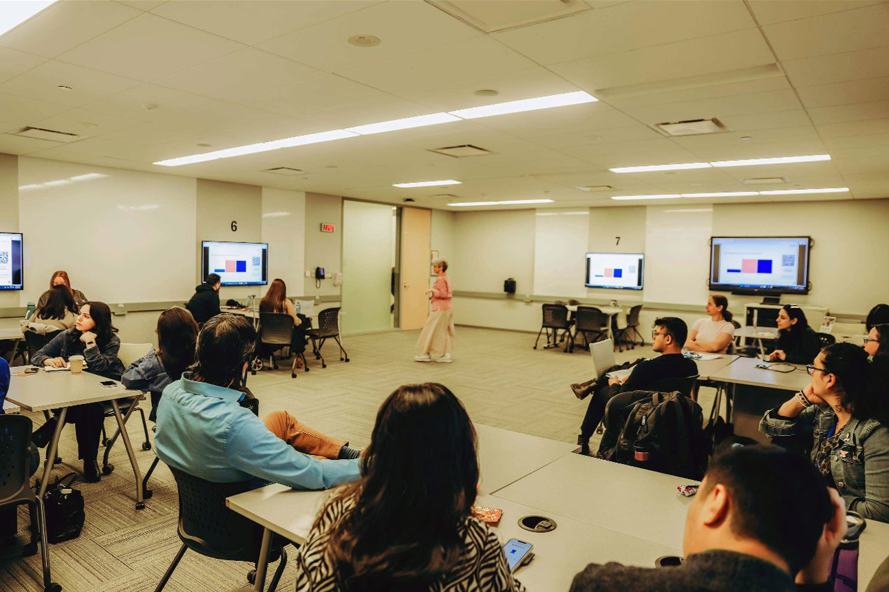 A modern classroom where an instructor stands at the front white adults seated at tables watch presentation screens or discuss the material in small groups.