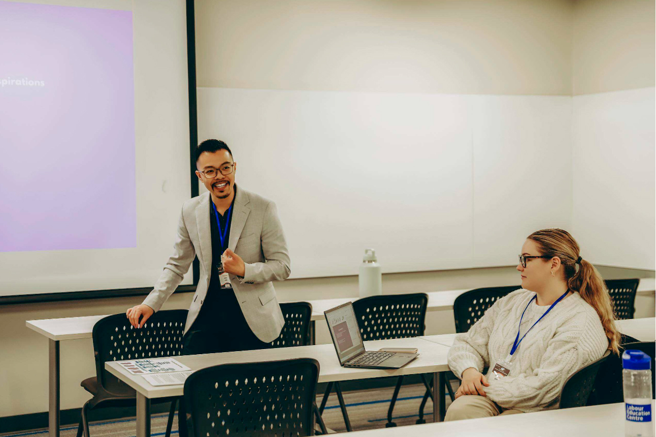 A facilitator wearing a light grey blazer stands at the front of a bright classroom, smiling and speaking to a small group during a presentation. A participant seated nearby listens attentively with an open laptop on the table. A projection screen, whiteboard, printed handouts, and reusable water bottles are visible, creating a collaborative professional learning environment.