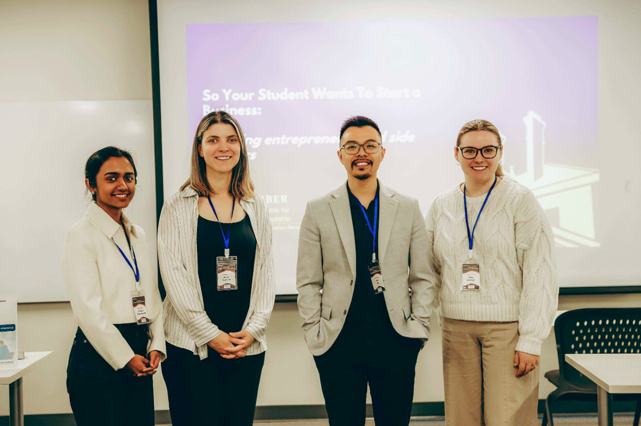 Four presenters stand side by side, smiling at the camera in a classroom after a presentation. Each person is wearing a conference name badge on a blue lanyard. Behind them, a projected presentation slide is displayed on a screen, and classroom tables and chairs are visible, highlighting a professional learning and collaboration setting.